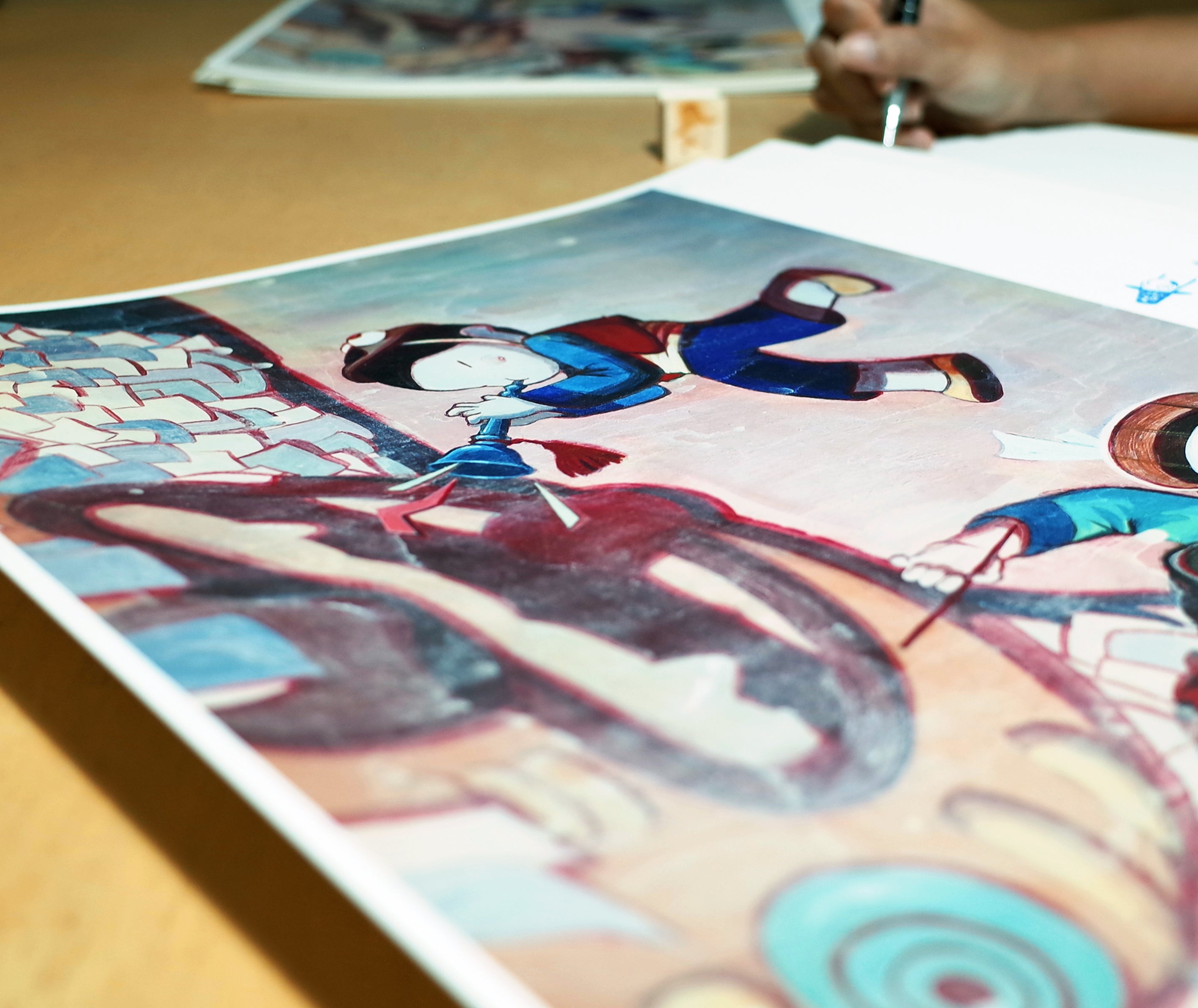 A person is creating a vibrant painting of animated characters, echoing Young-Ji Chas Festival (마당놀이). A close-up shows the artists hand with a pen, hinting that this potential archival pigment print is still in progress.