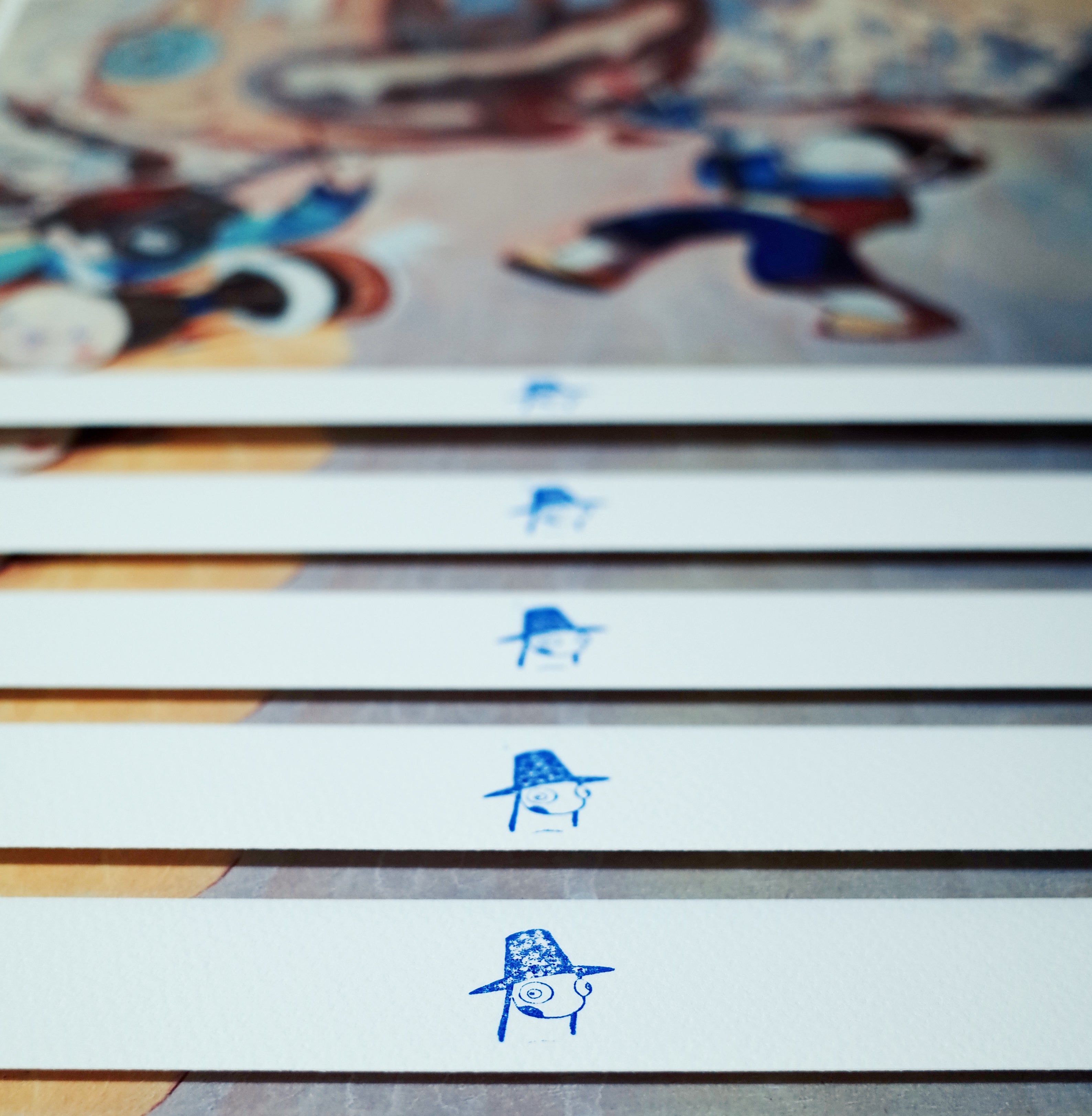 Close-up of several sheets of Young-Ji Chas Festival (마당놀이) prints, each showcasing a small blue stylized face with a hat like festival masks, meticulously aligned with blurred colorful artwork in the background.