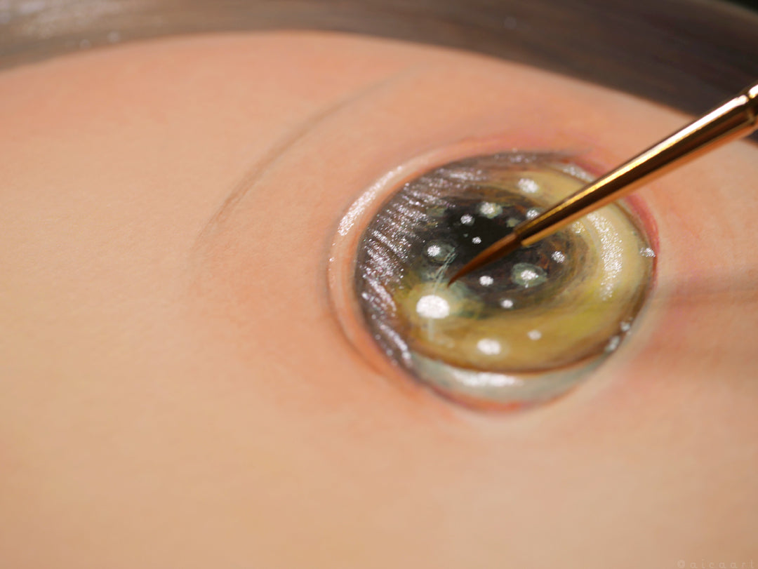 A close-up shows a hand using a fine paintbrush, skillfully hand embellishing Heart by aica. The shimmering reflections and intricate colors in this realistic eye illustration are destined for signed and numbered archival pigment print masterpieces.