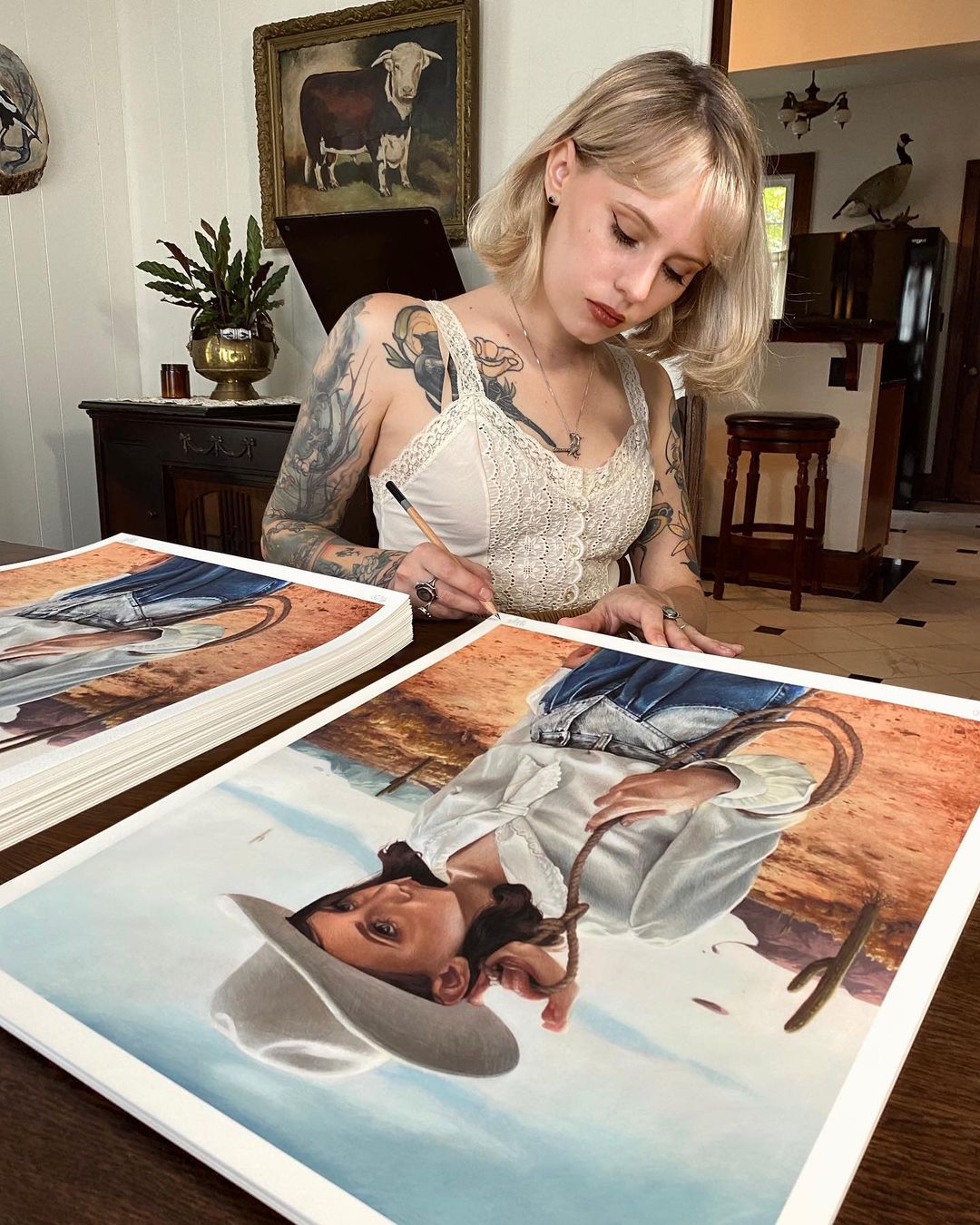 A blonde woman with floral tattoos sits at a table, signing Alone With My Thoughts archival pigment prints by Meagan Magpie Rodgers, featuring a person in a cowboy hat. She wears a lace top, surrounded by western-themed decor like paintings and a plant.