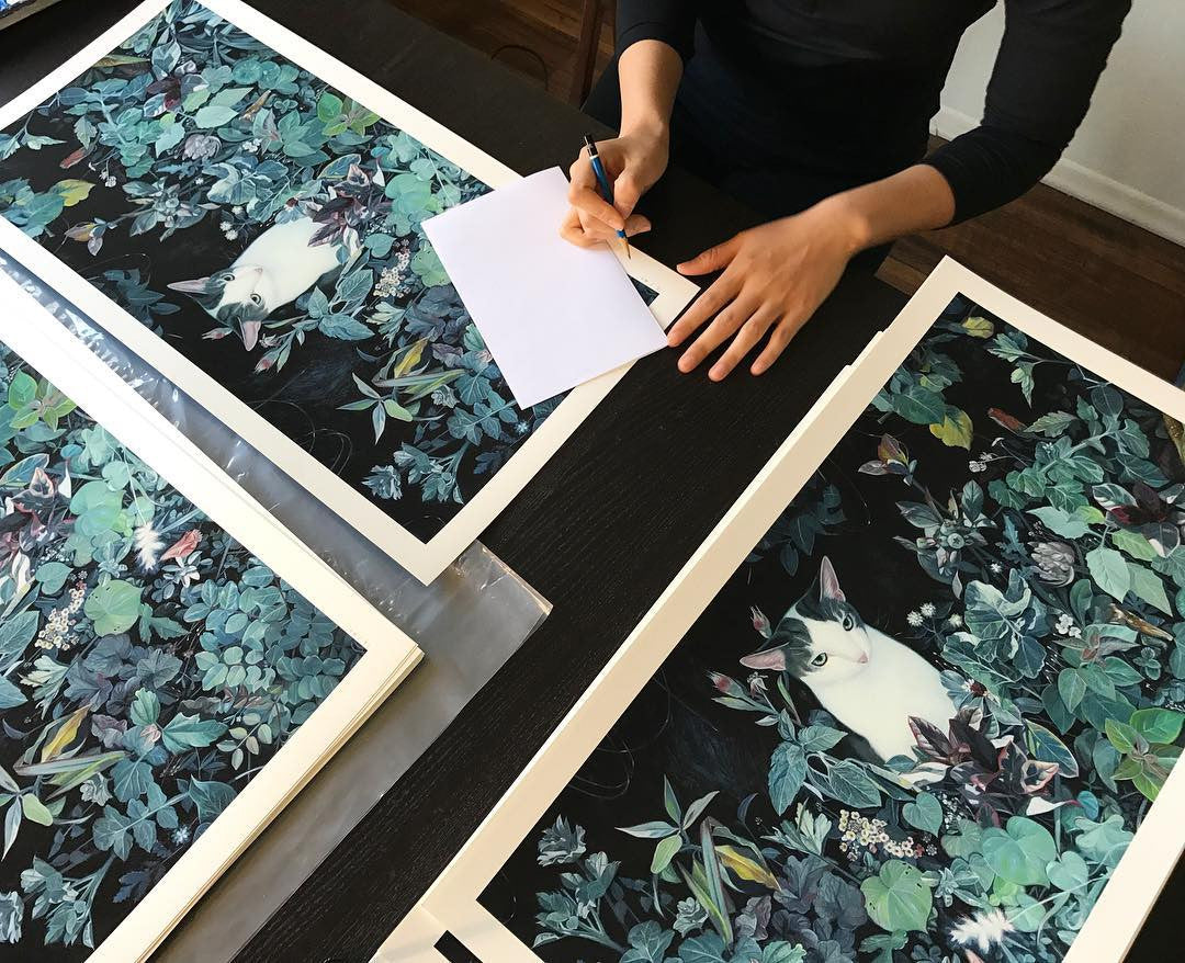 Joanne Nams A Quiet Soul limited edition art prints depict a cat amid dense foliage, with green and blue leaves. These archival pigment prints, hand embellished for depth, are spread on a dark table as the person gracefully works with pen and paper.