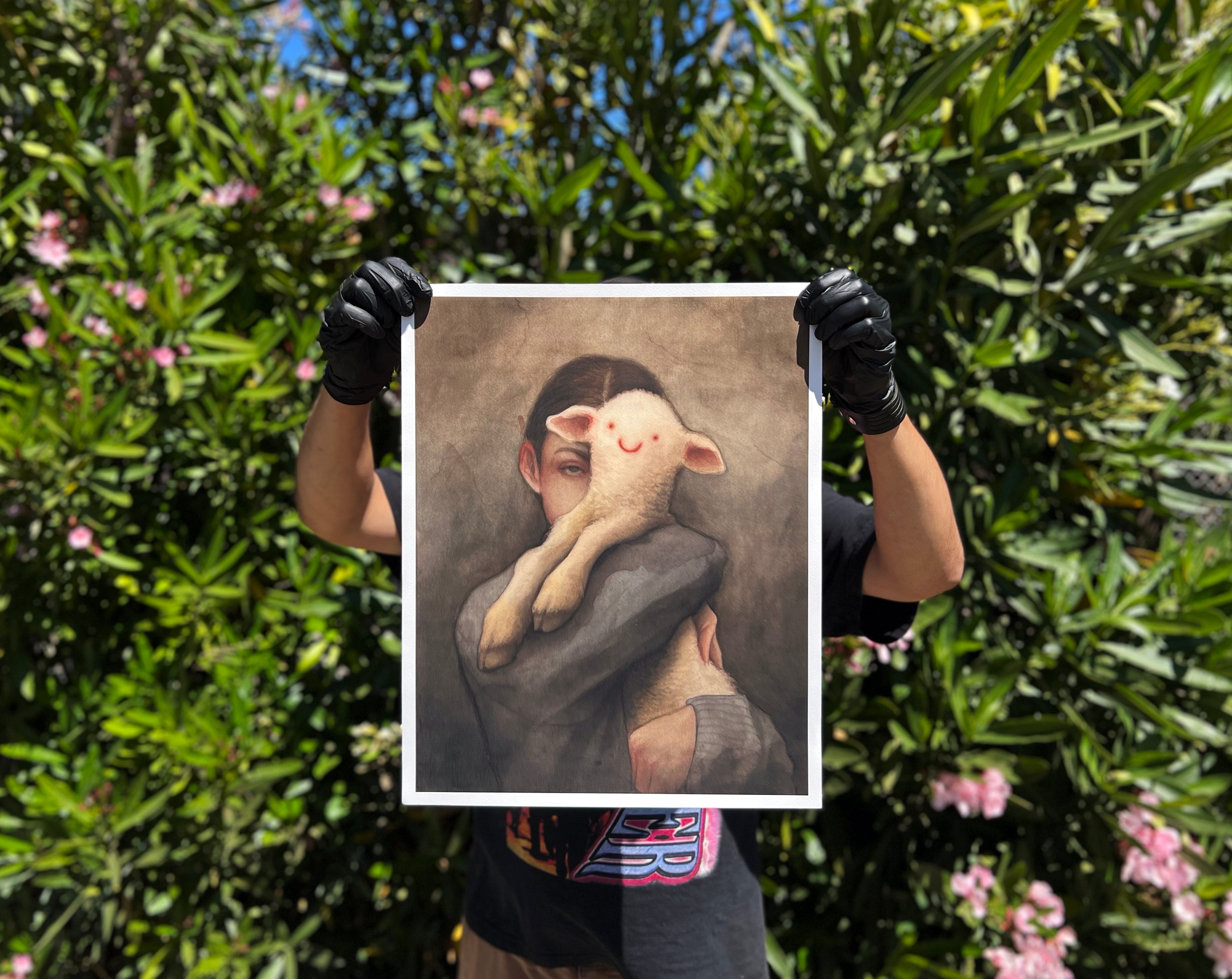 A person in black gloves holds a Lamb Archival Pigment Print by Robert Duxbury, depicting a smiling lamb in someones arms, set against lush greenery with blooming flowers. Includes Certificate of Authenticity.
