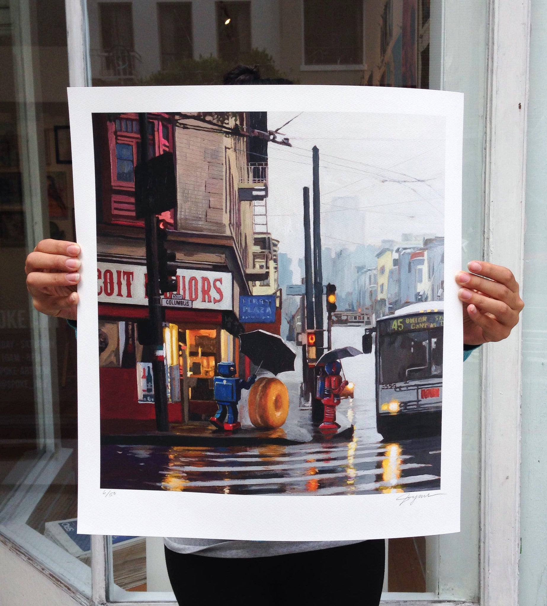 A person holds the limited edition North Beach by Eric Joyner, showcasing a rainy urban street scene with a liquor store, people with umbrellas, a fire hydrant, doughnut sculpture, and bus 45. Reflections shimmer on the wet pavement in this fine art piece.