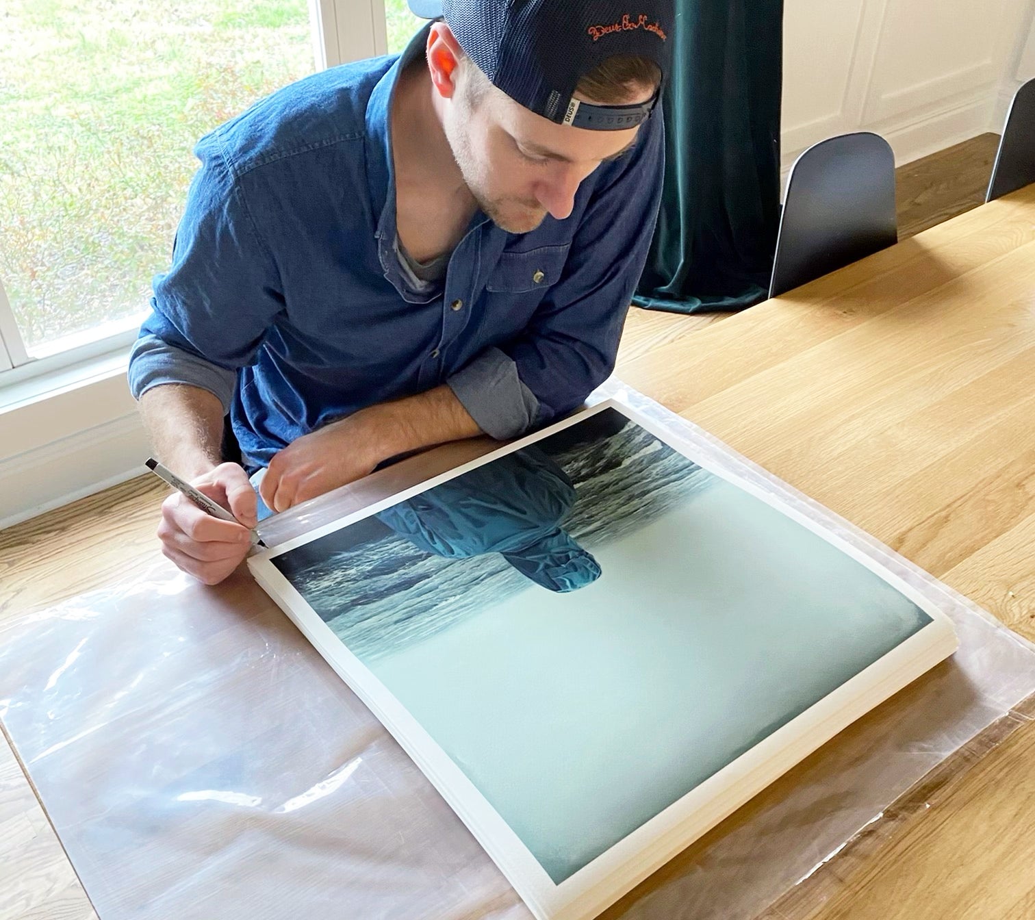 At a wooden table, a person in a blue shirt and cap signs and numbers Feel It Running Down My Spine, an archival pigment print by Adam Hall. The limited edition features someone in a blue jacket in a misty landscape.