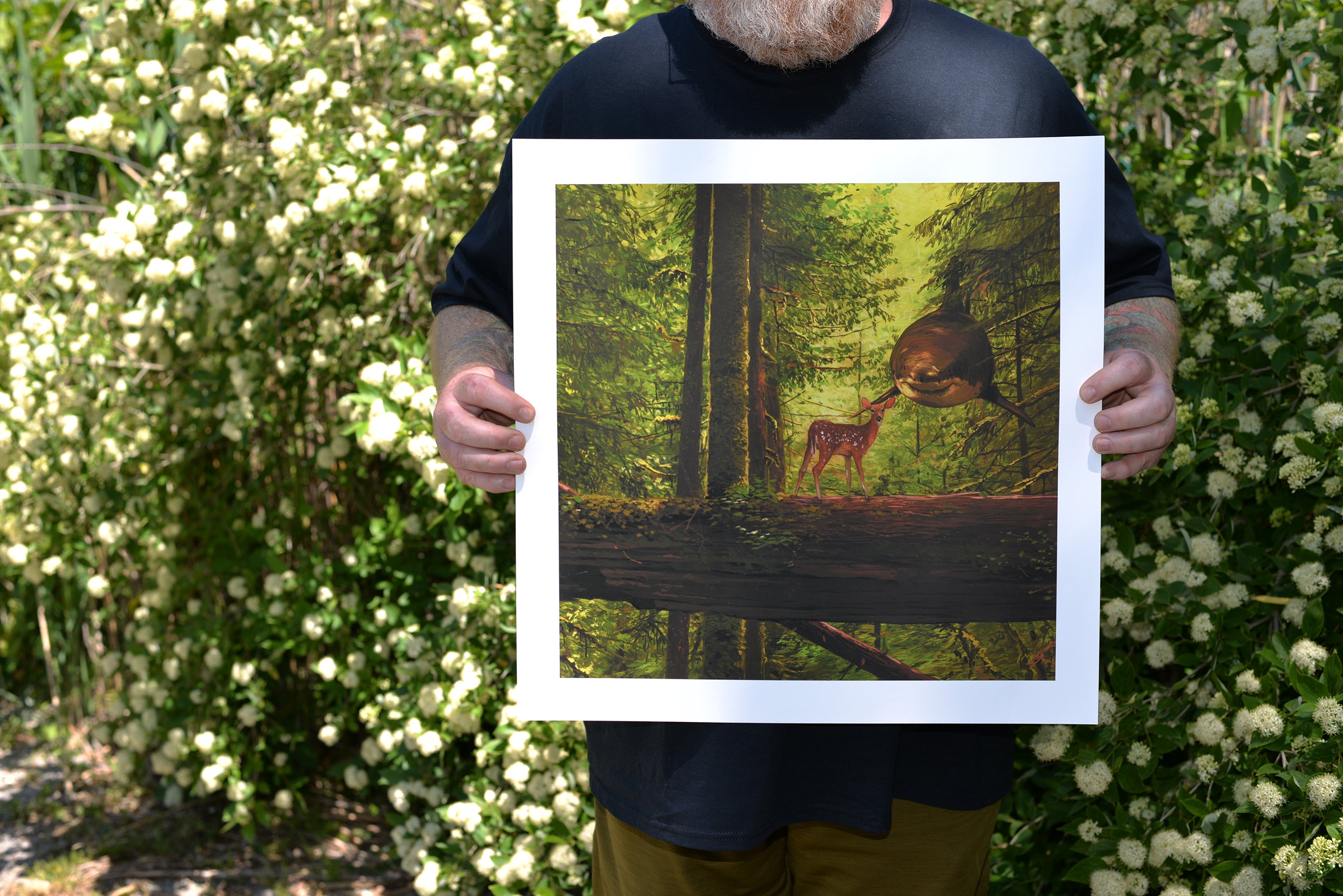 A person holds You Cried Out To Me, a limited edition archival pigment print by Chris Austin, depicting a forest scene with a deer and reflective orb. Standing near a flowering shrub, they wear a black shirt and brown pants, their face unseen.