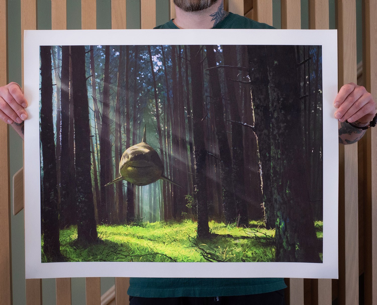 Someone holds a Chris Austin Ill Hear You Calling Me poster depicting a surreal scene of a large, smiling fish in a sunlit forest. Sunrays illuminate both the fish and vibrant foliage. This archival pigment print is signed and numbered for authenticity.