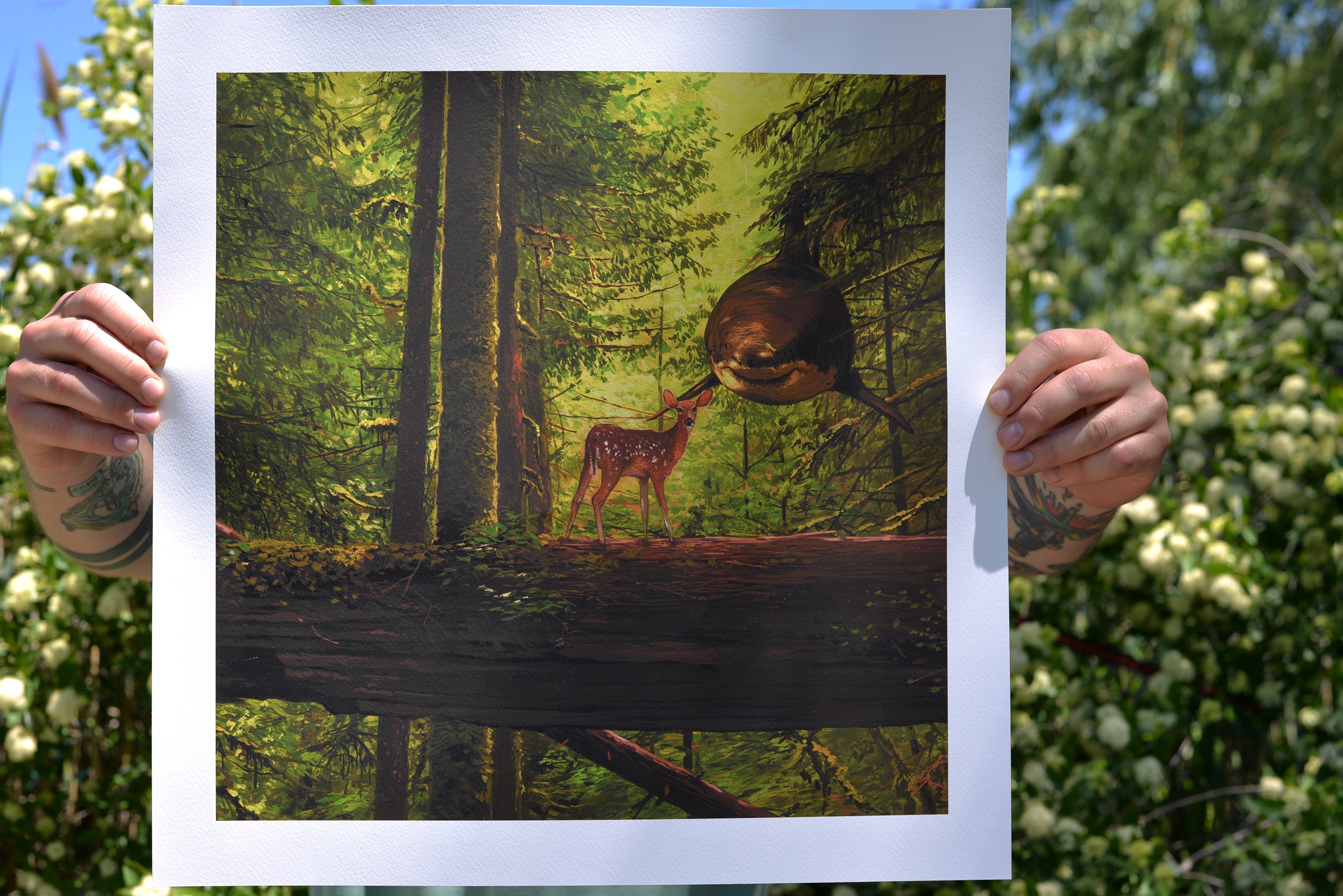 A person holds You Cried Out To Me, a limited edition archival pigment print by Chris Austin, depicting a fawn and large bird on a log surrounded by lush green trees with flowering bushes in sunlight.