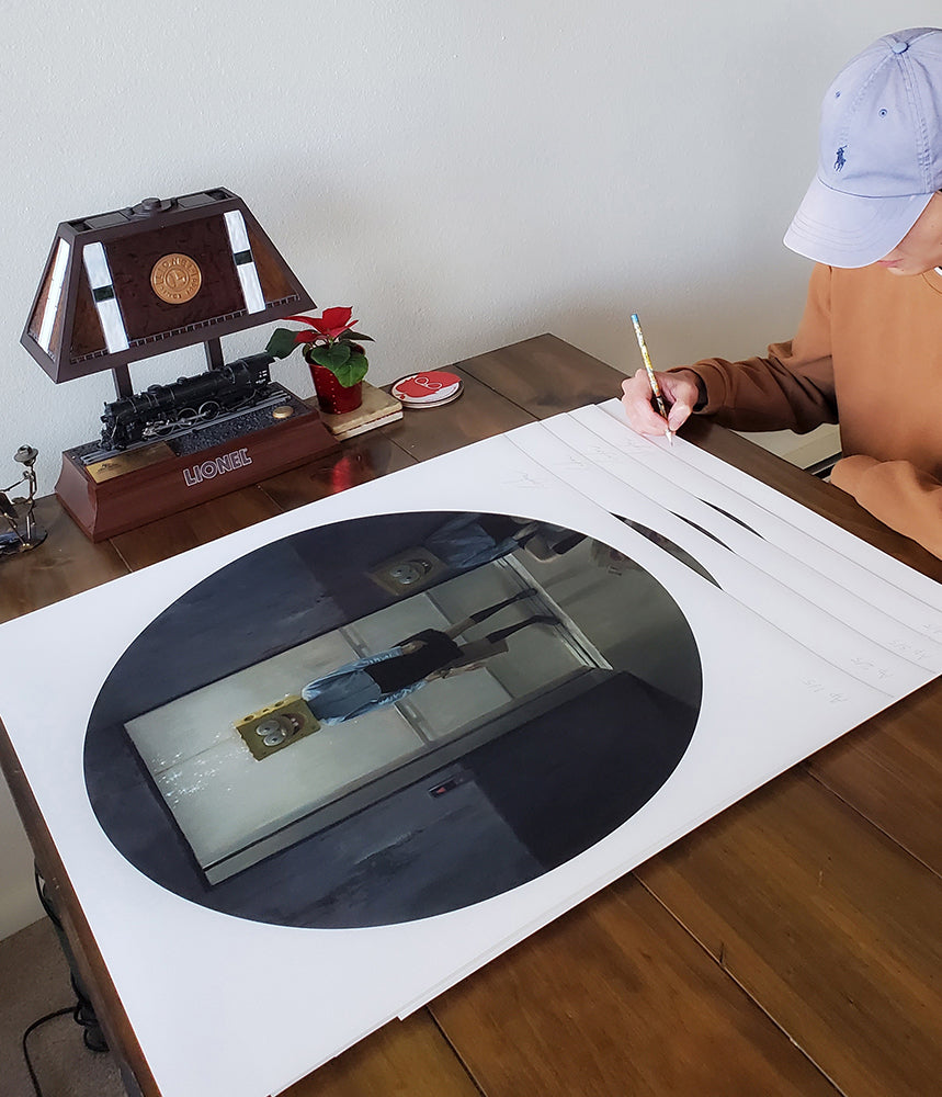 A person in a hat sketches on a large white paper featuring Time Traveler, a black oval by Leegan Koo, depicting a reflective hallway. An archival pigment print enhances the scene with a model train lamp and small plant on the wooden table.