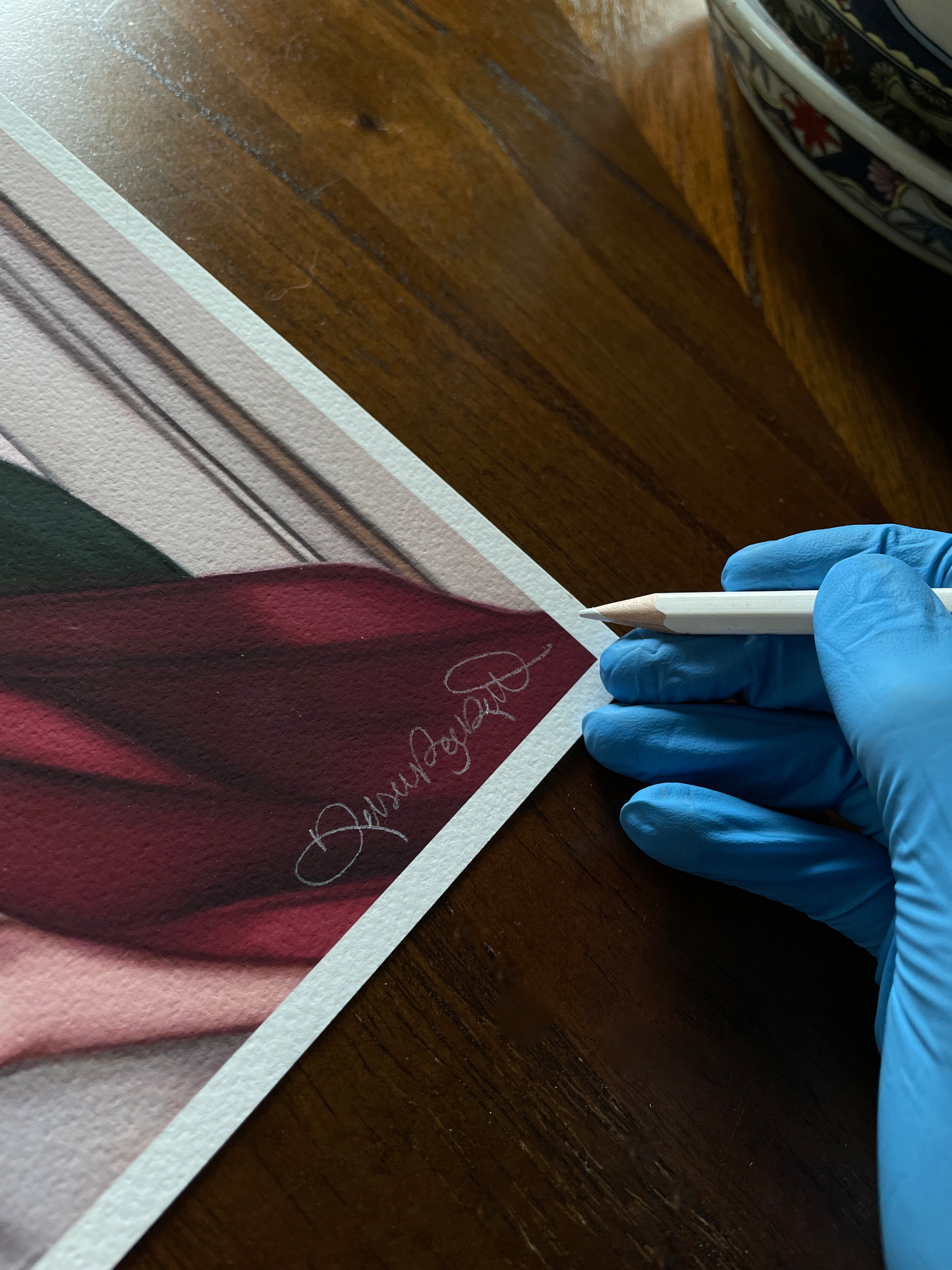 Wearing blue gloves, someone carefully signs the corner of Another Last Time, an Archival Pigment Print by Kelsey Beckett, with a pencil. The print features rich maroon and dark tones on a wooden table, alongside a decorative bowl in the background.
