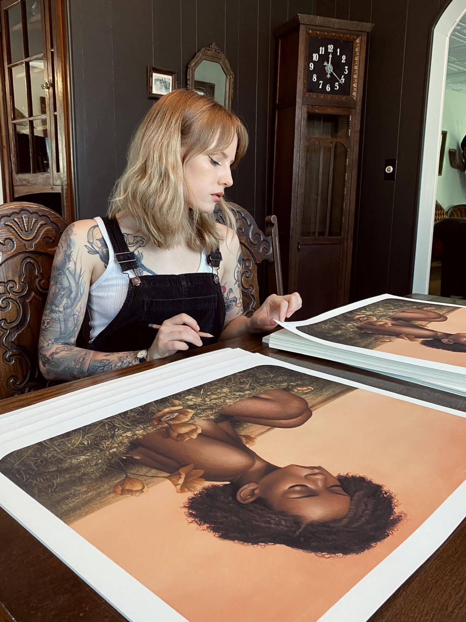 A tattooed individual sits at a table, studying Efflorescence archival pigment prints by Meagan Magpie Rodgers, featuring a child with curly hair and animals on an orange backdrop. A vintage clock and wooden furniture enhance this vibrant scene.