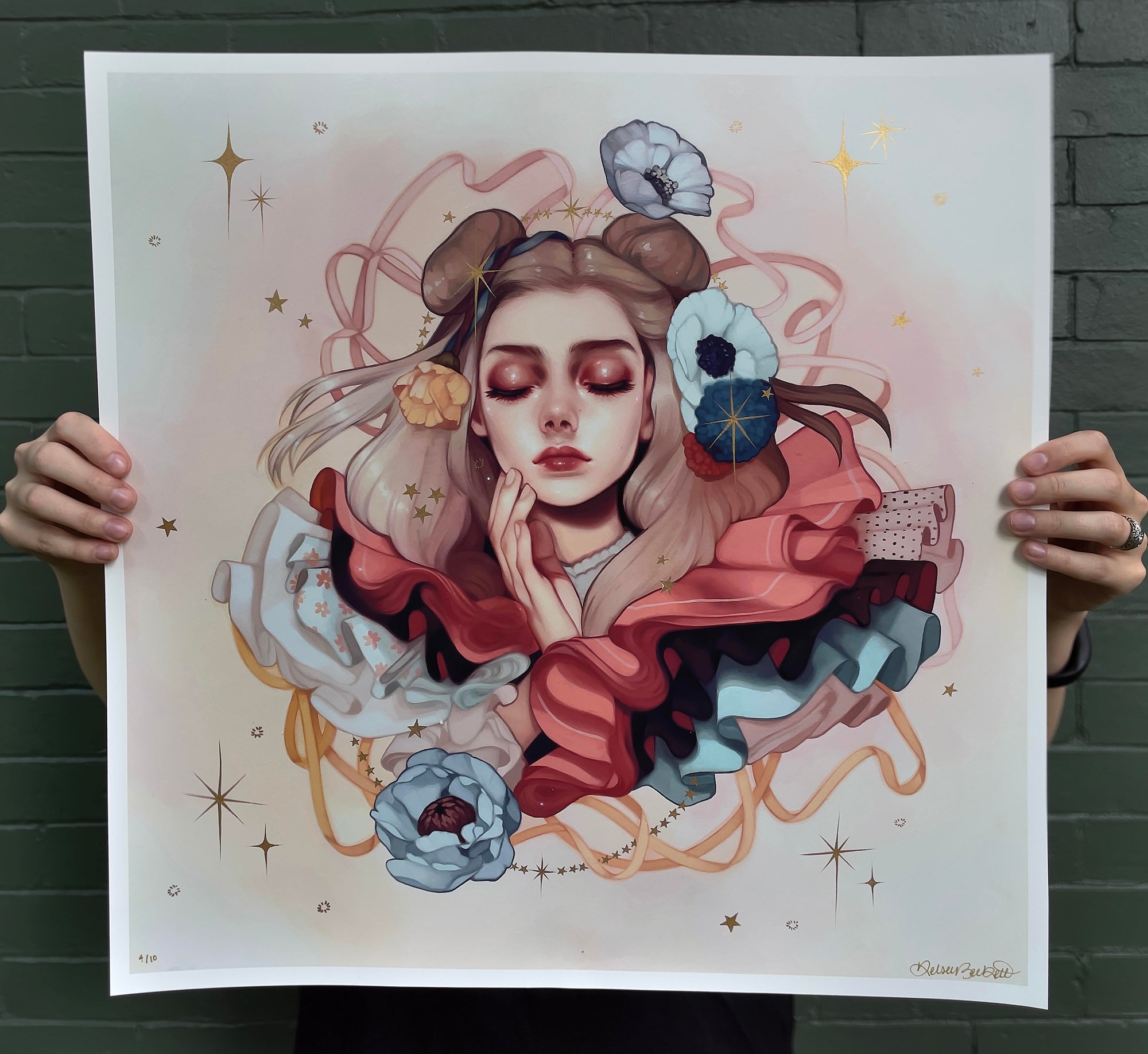A person holds Kelsey Becketts Abundance, a colorful archival pigment print of a woman with closed eyes, surrounded by abstract swirls, flowers, and stars. Hand embellished with bold red, blue, and gold hues, it creates a whimsical and dreamy atmosphere.