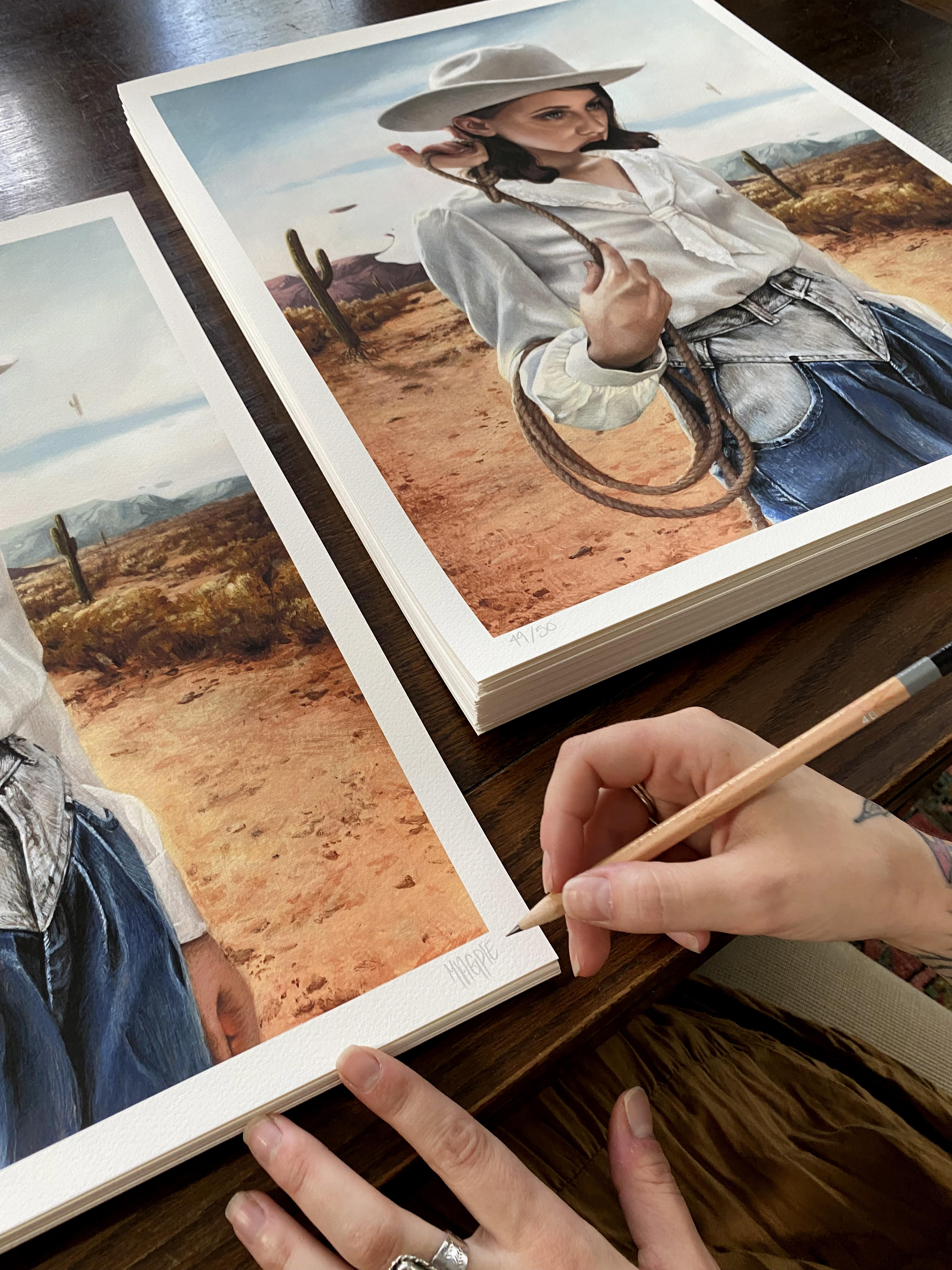 Artist Meagan Magpie Rodgers signs archival pigment prints, Alone With My Thoughts, depicting a woman in a western outfit amidst cacti and desert. The print, featuring hands with rings holding a pencil, rests on a dark wooden table.