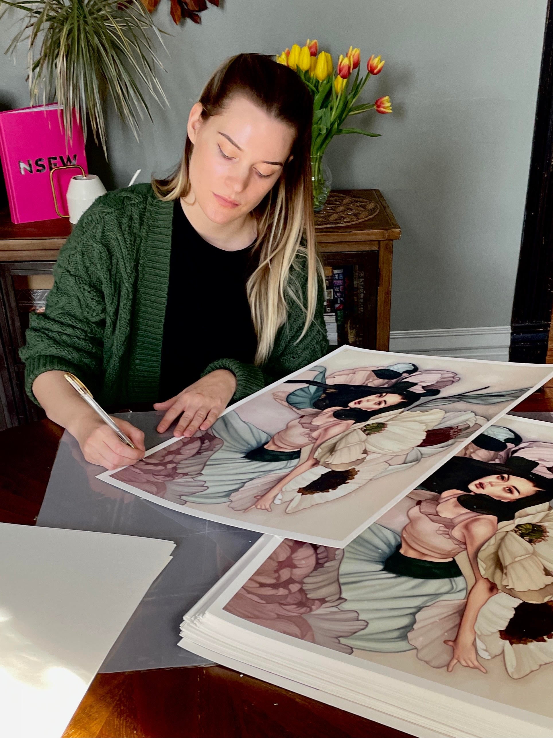 Artist Kelsey Beckett, wearing a green cardigan, elegantly signs her archival pigment prints titled The Flower Girl while sitting at a wooden table adorned with tulips and a pink book.