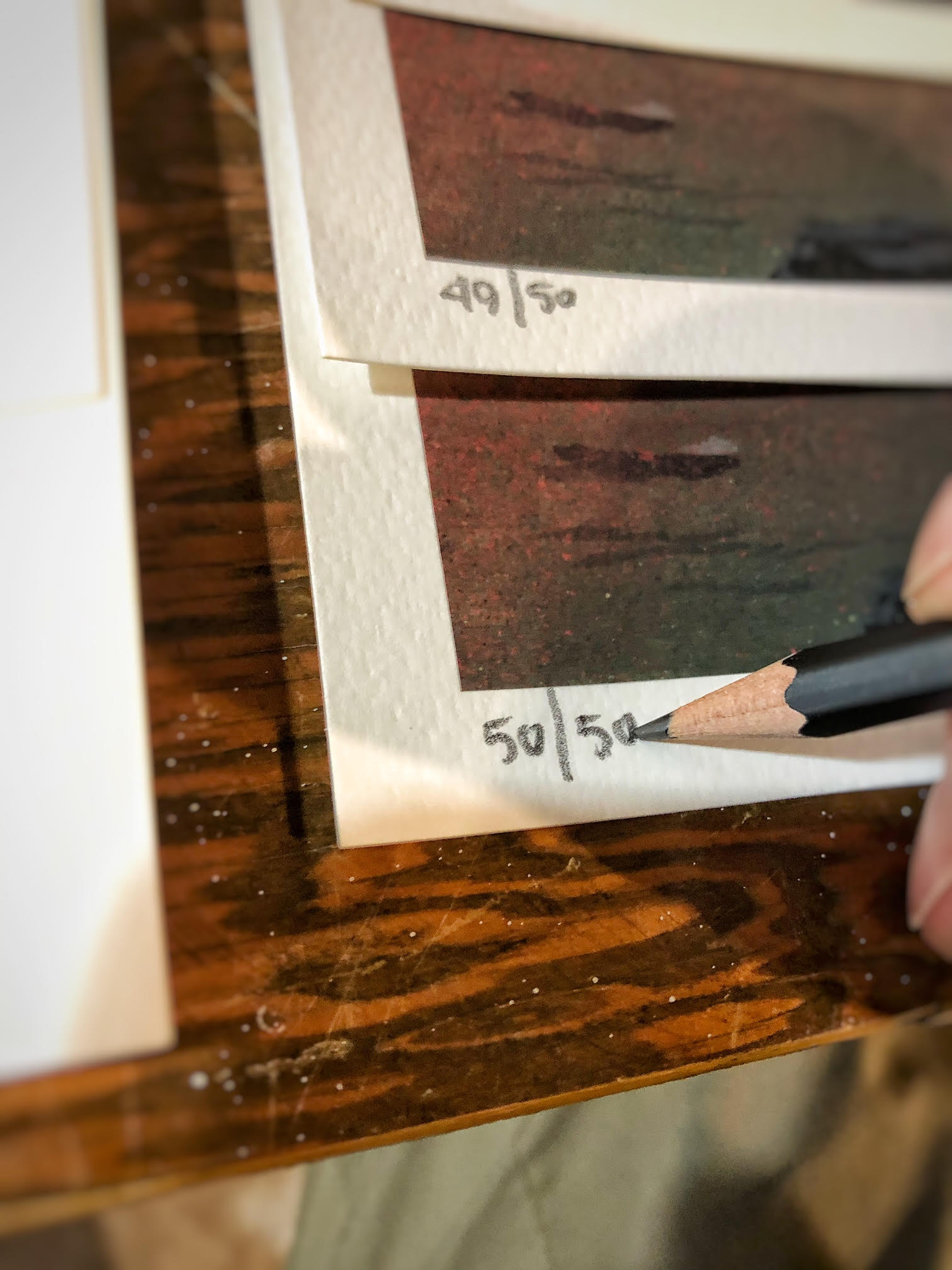 A close-up shows a hand holding a black pencil, inscribing 50/50 onto textured paper—likely part of Chris Austins Art Edition, In The Teeth Of A Crashing Gale. Below it, a partially hidden sheet marked 49/50 rests on the wooden surface like an archival pigment print.
