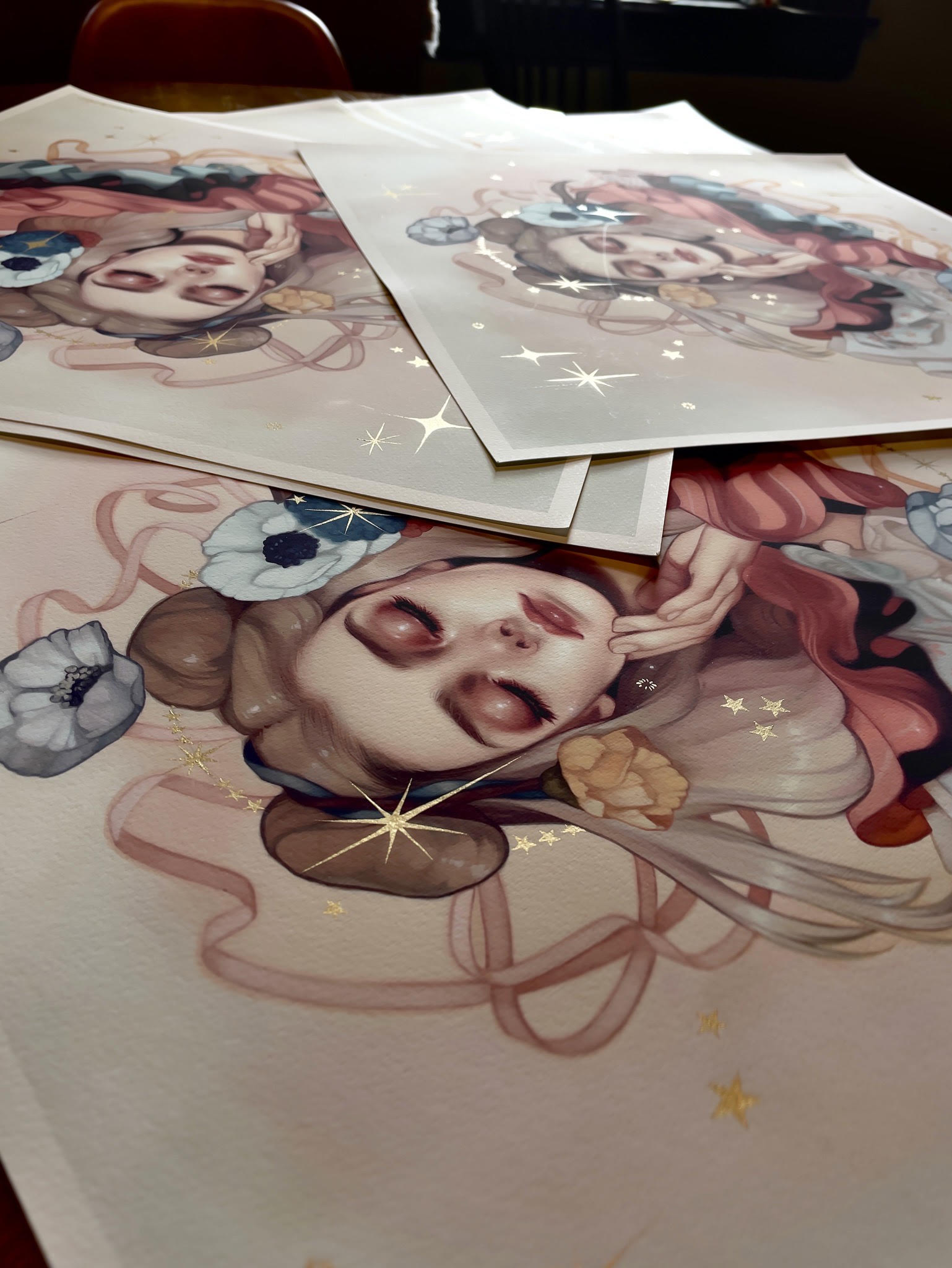 The Abundance art print by Kelsey Beckett features a stylized portrait with closed eyes surrounded by flowers and ribbons. This Hand Embellished Edition uses a soft palette with gold star accents, creating a sense of abundance.