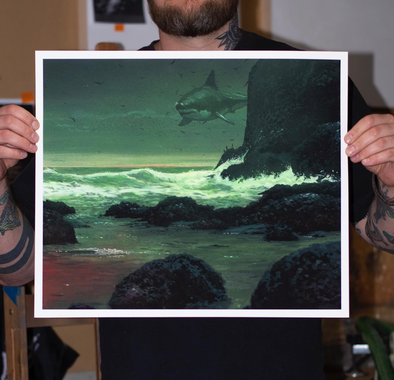 A tattooed individual holds Chris Austins In The Teeth Of A Crashing Gale, featuring a dramatic scene with a large shark hovering above an illuminated, rocky sea beneath a stormy sky, blending realistic waves with an archival pigment print of the surreal creature.