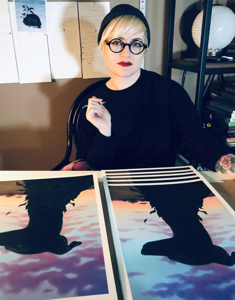 A person in glasses and a beanie sits at a table with the Bitter Blooms limited edition artwork by Syd Bee, showing crows against a vibrant sky. Holding a pen amidst shelves and pinned papers, theyre possibly preparing another signed and numbered masterpiece.
