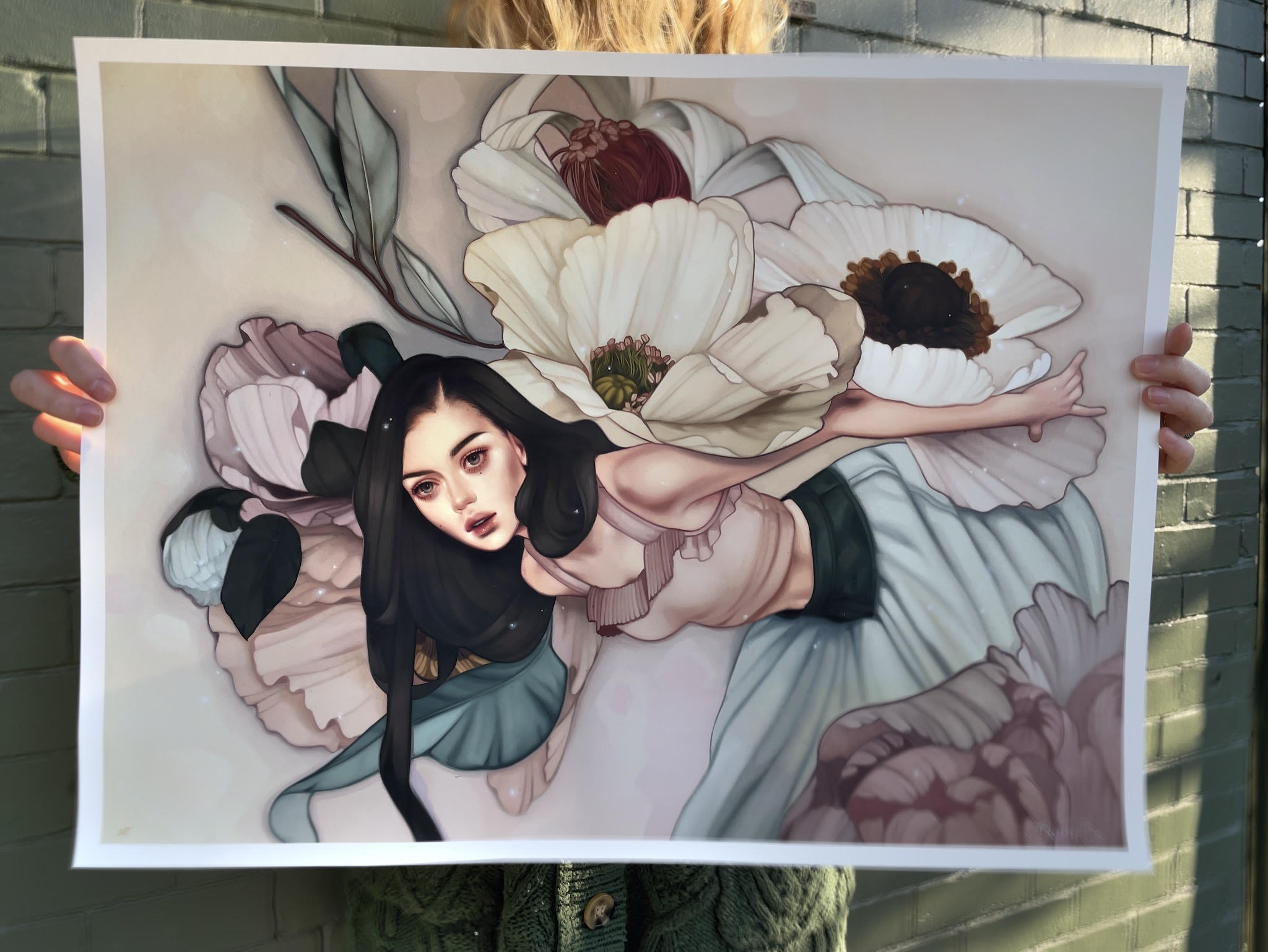 A person holds a large archival pigment print, The Flower Girl by Kelsey Beckett, showing a woman with long dark hair and giant white and pink flowers. The whimsical figure has outstretched arms, a peaceful expression, and blends into the floral background.