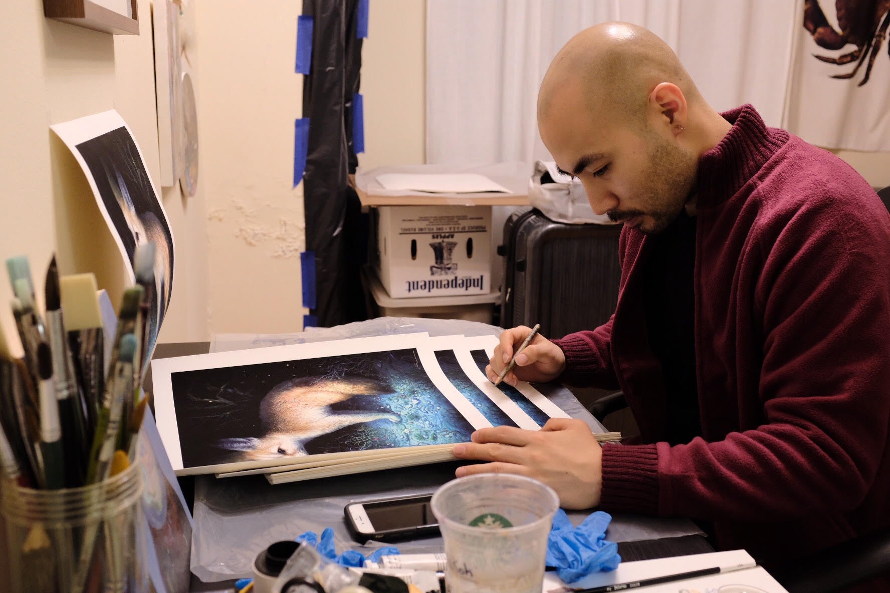 In a studio setting, a bald person in a maroon sweater hand embellishes and signs a large Neith print by KiSung Koh. Art supplies such as brushes and a cup surround them as they focus on the numbered edition artwork featuring a fox.