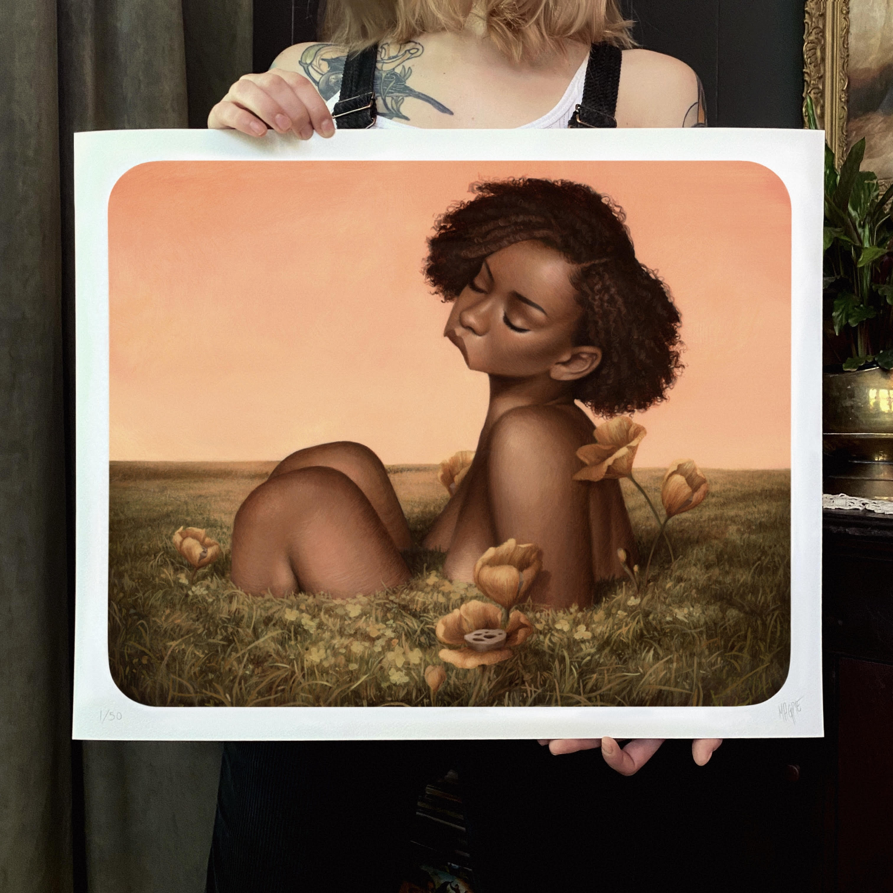 Someone holds a framed archival pigment print titled Efflorescence by Meagan Magpie Rodgers, showcasing a serene figure blending with a grassy landscape and orange flowers under a warm sky, embodying the harmony of human form and natures tranquility.